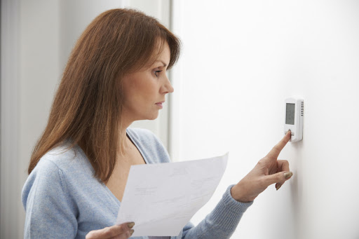A woman holding a bill as she adjusts the thermostat in a home.