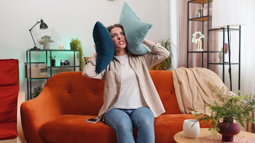 A woman sitting on a couch and covering her ears with pillows due to a loud noise.