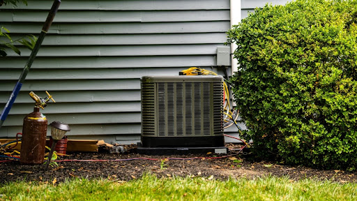 A central AC unit installed outside of a house near a bush.