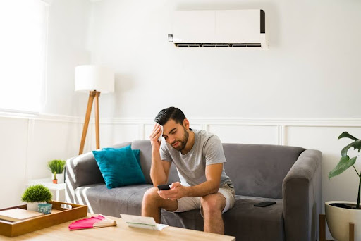 Man sitting on couch in living room wiping sweat off his brow because of broken AC.